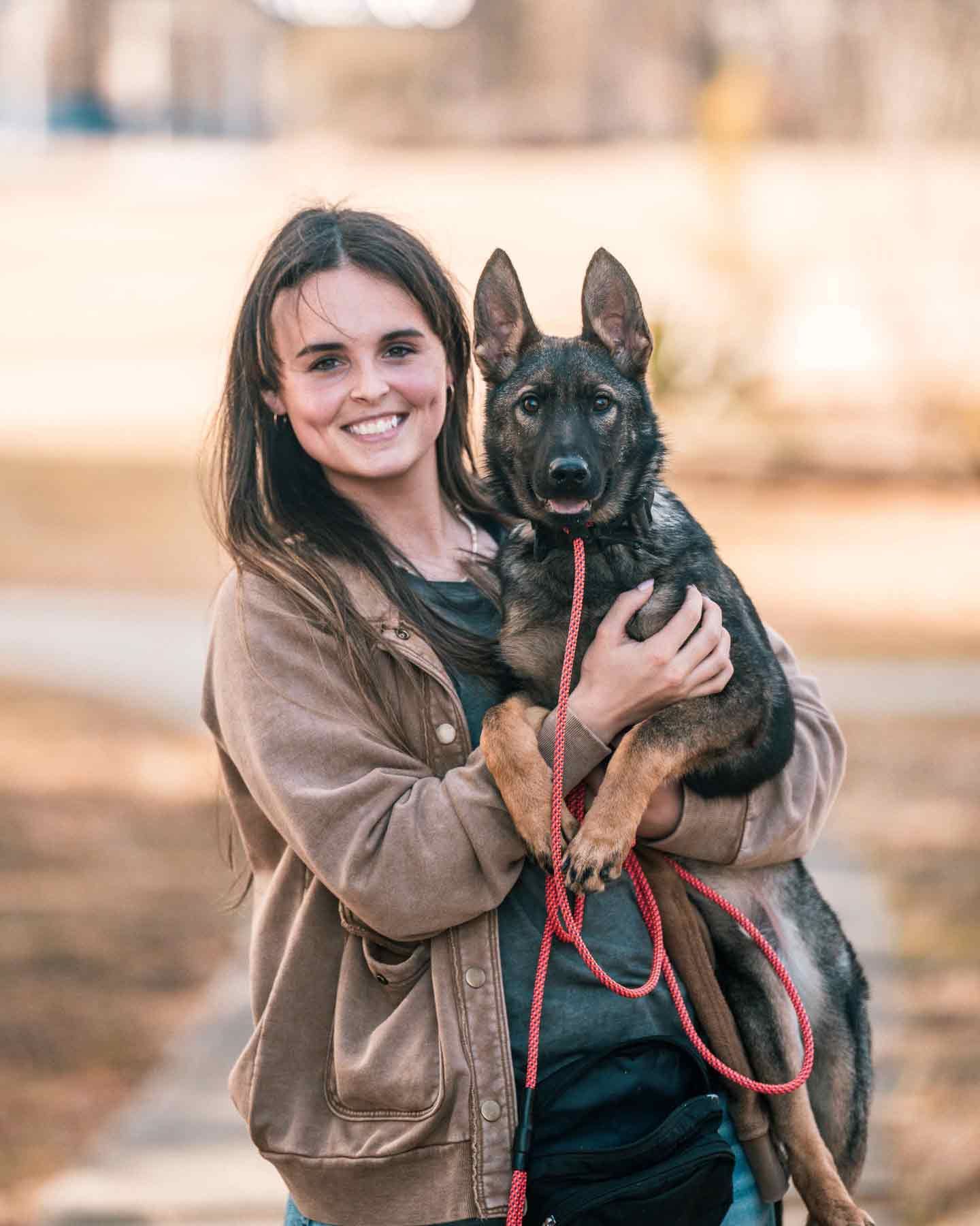 Woman smiling, holding a German Shepherd puppy outdoors; brown coat, leash, blurred background.