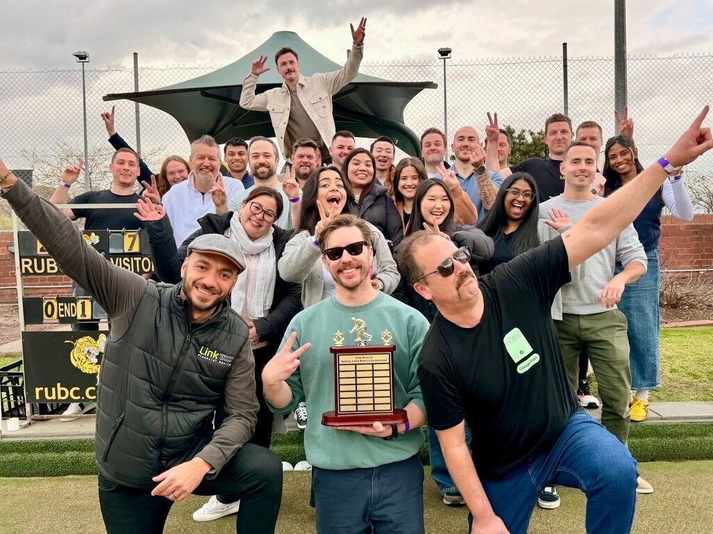 Group of people celebrating outdoors, holding trophy. One person raises arms, others smile. A golf course is the background.