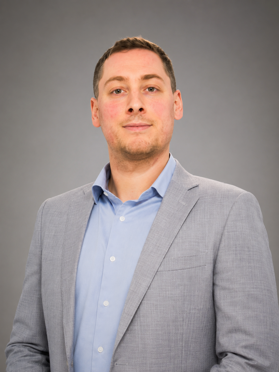 Man in a gray blazer and light blue shirt stands with hands clasped in front of a wood-paneled wall.