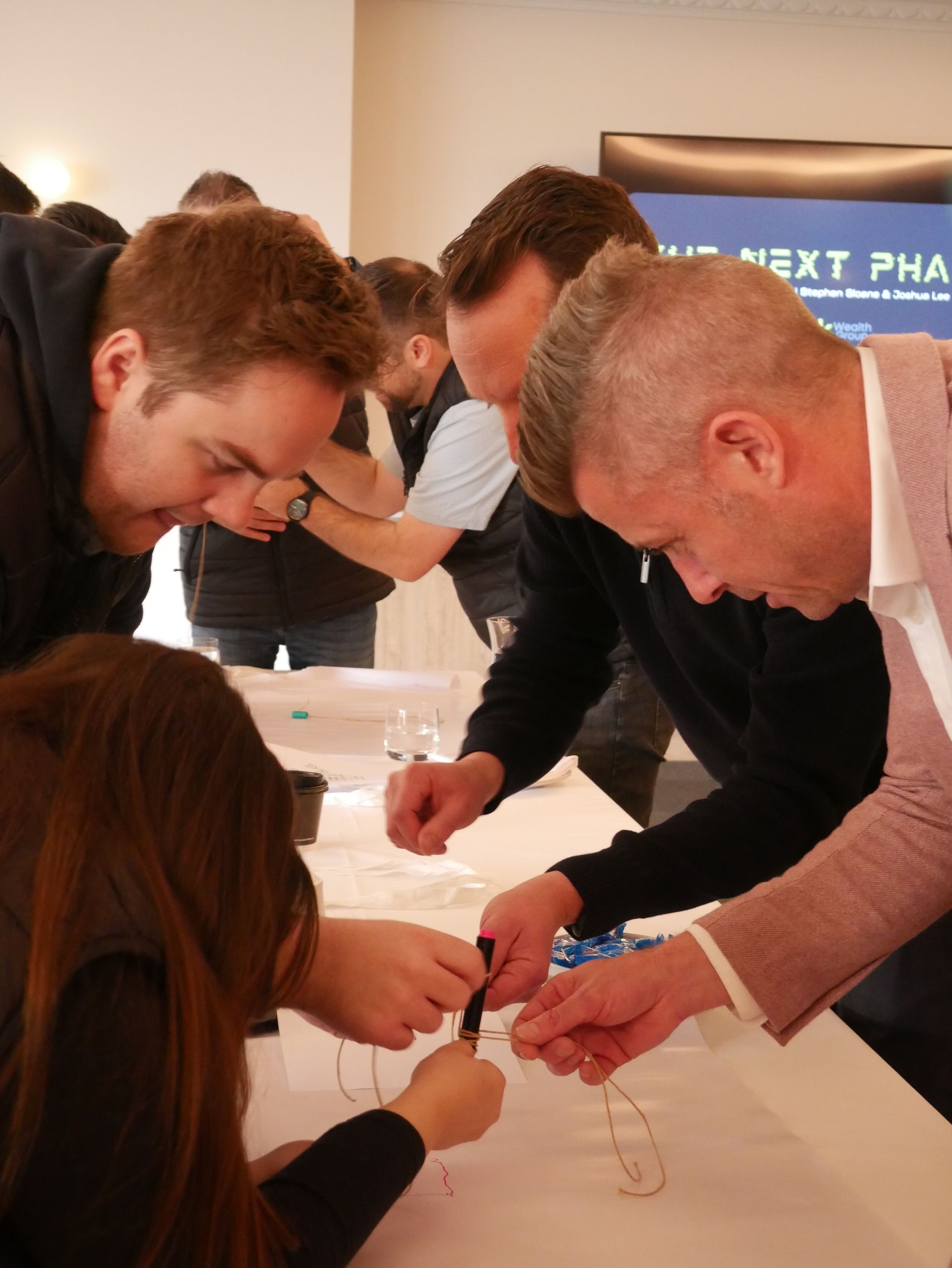 People leaning over a table, focused on manipulating wires or small objects; indoor setting.