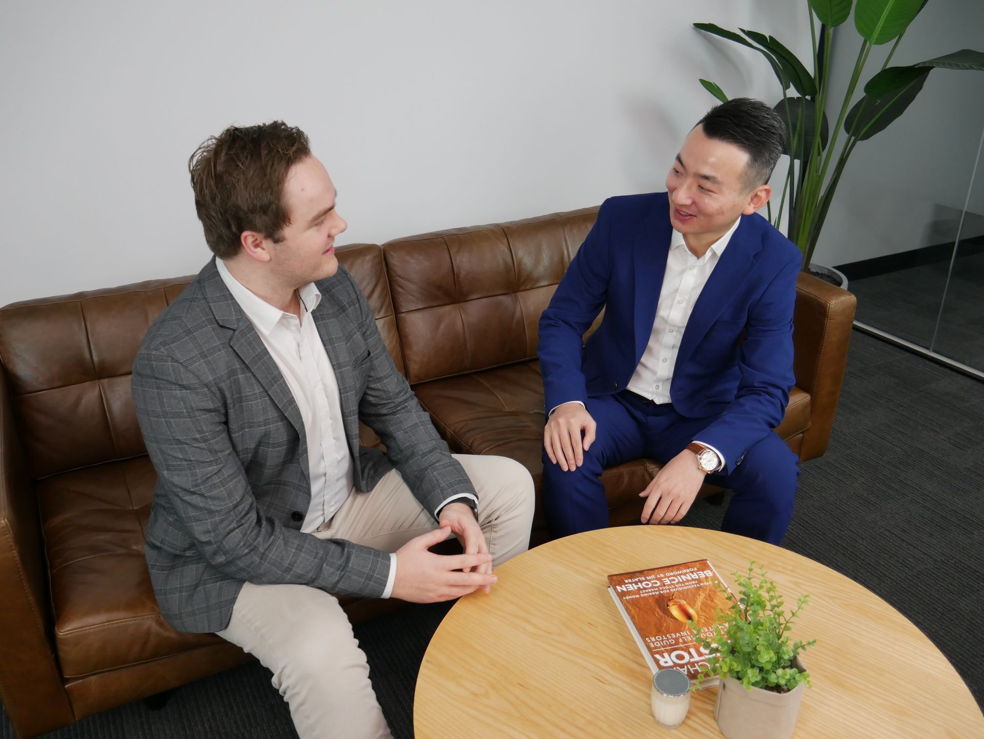 Two men shaking hands on a brown leather couch; one in a tan blazer, one in a light blue jacket.