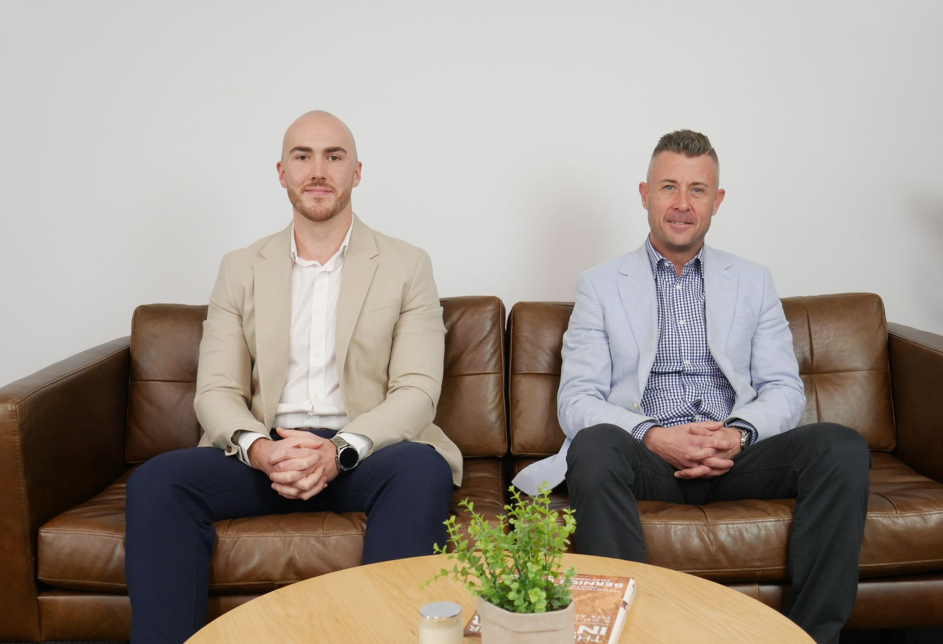 Two men seated on a brown sofa, in business attire, looking at the camera. A small plant on a table in front.