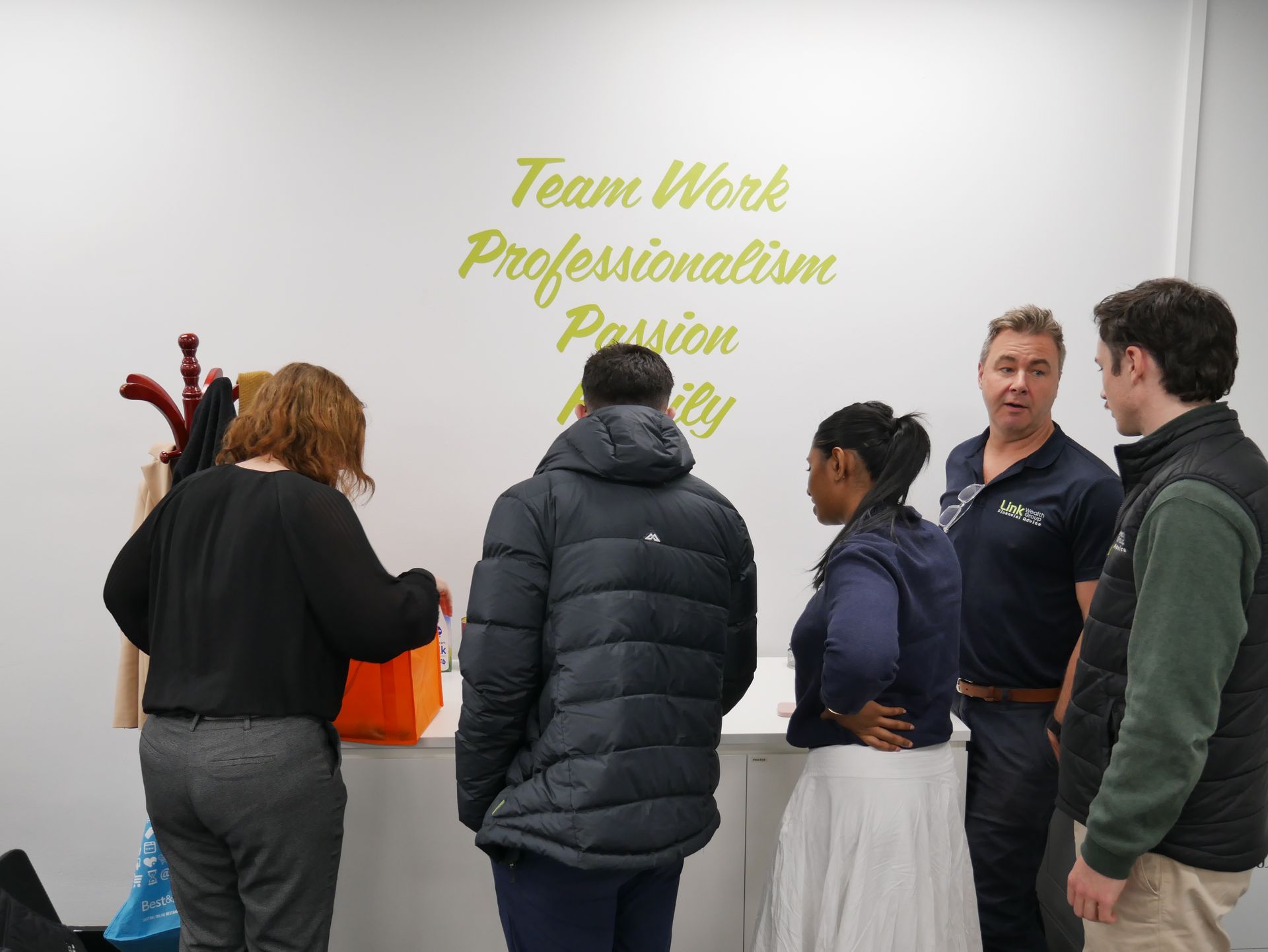 Group of people near a white wall with company values written in gold. One person is opening a gift bag.