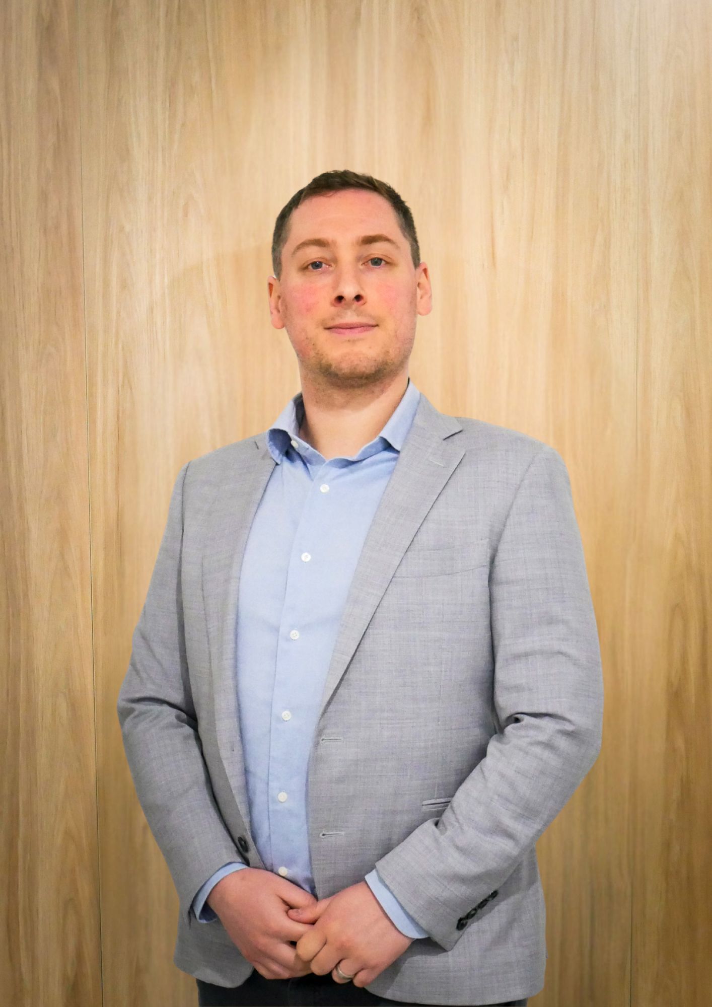 Man in a gray blazer and light blue shirt stands with hands clasped in front of a wood-paneled wall.
