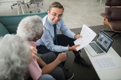Man in a suit showing documents to an older couple seated together; laptop and papers on table.