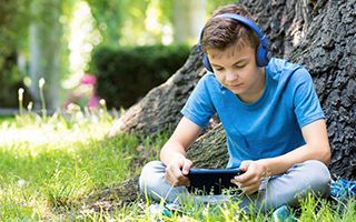 A young boy wearing headphones is sitting under a tree using a tablet.