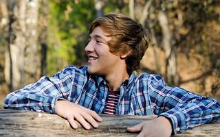 A young man in a plaid shirt is leaning on a wooden fence.