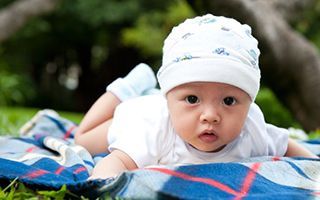 A baby wearing a hat is laying on a blanket in the grass.