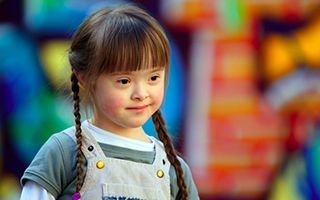 A little girl with down syndrome is smiling in front of a colorful background.