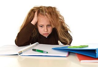 A little girl is sitting at a desk with her hands on her head.