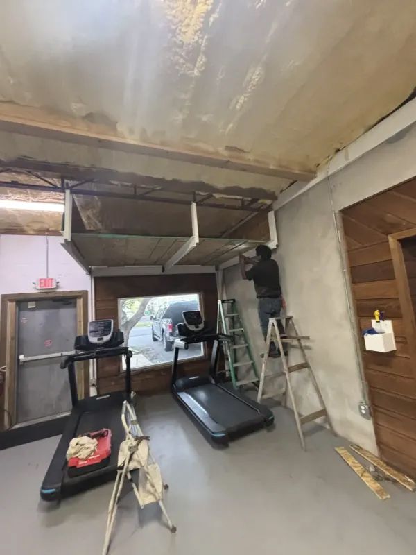 Man on ladder working on ceiling structure in gym with treadmills.