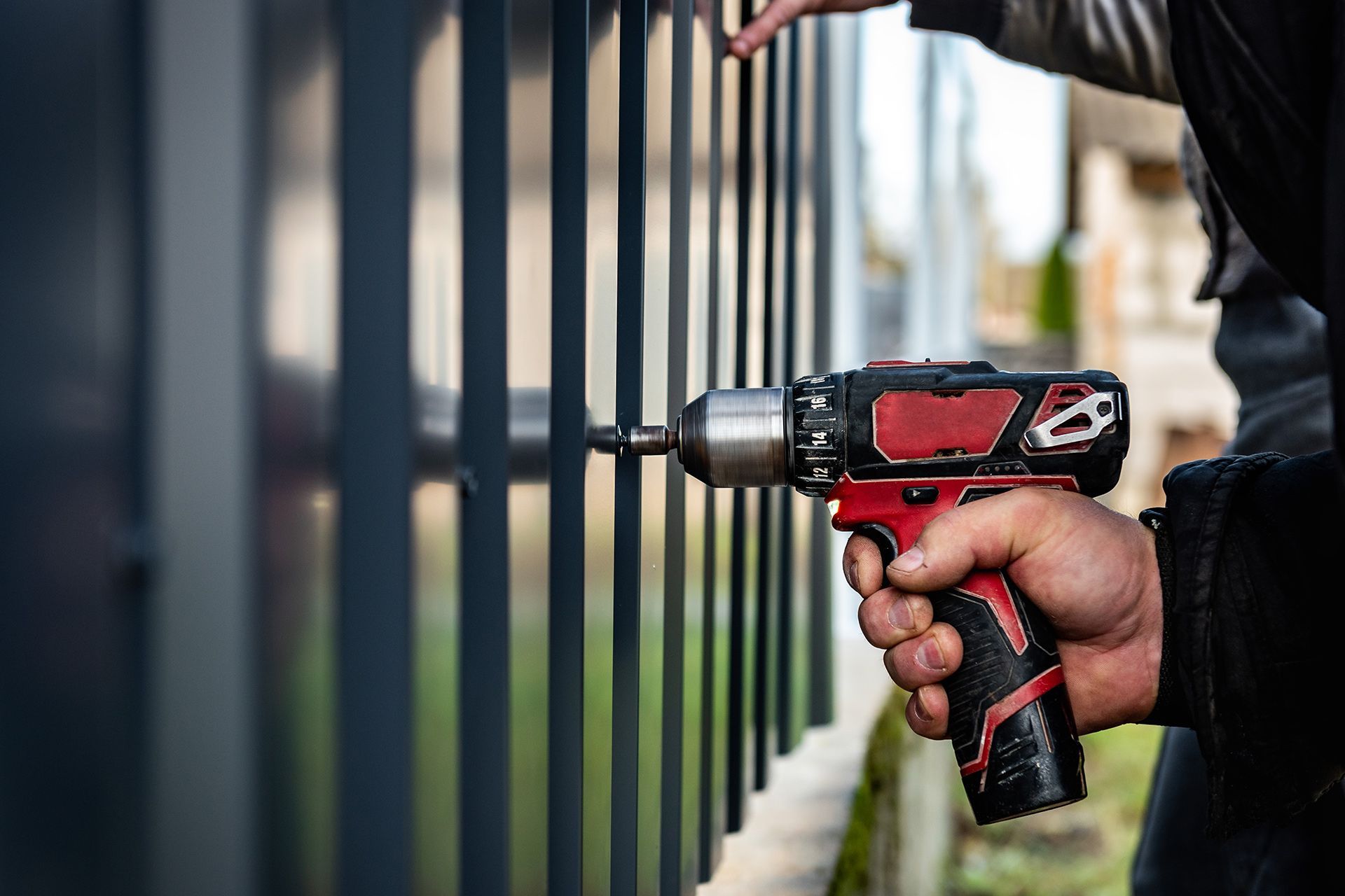Person using a red and black power drill to install a dark gray metal fence.