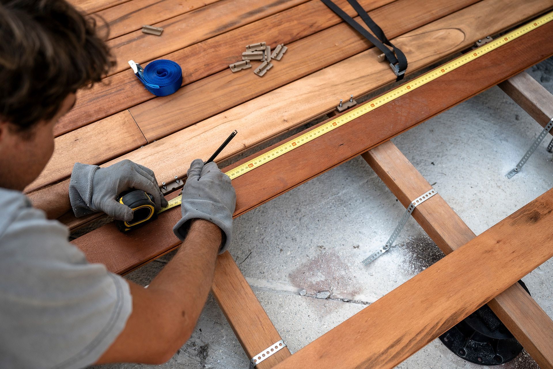 Person in work gloves measuring and marking wooden deck planks with a measuring tape and pencil.