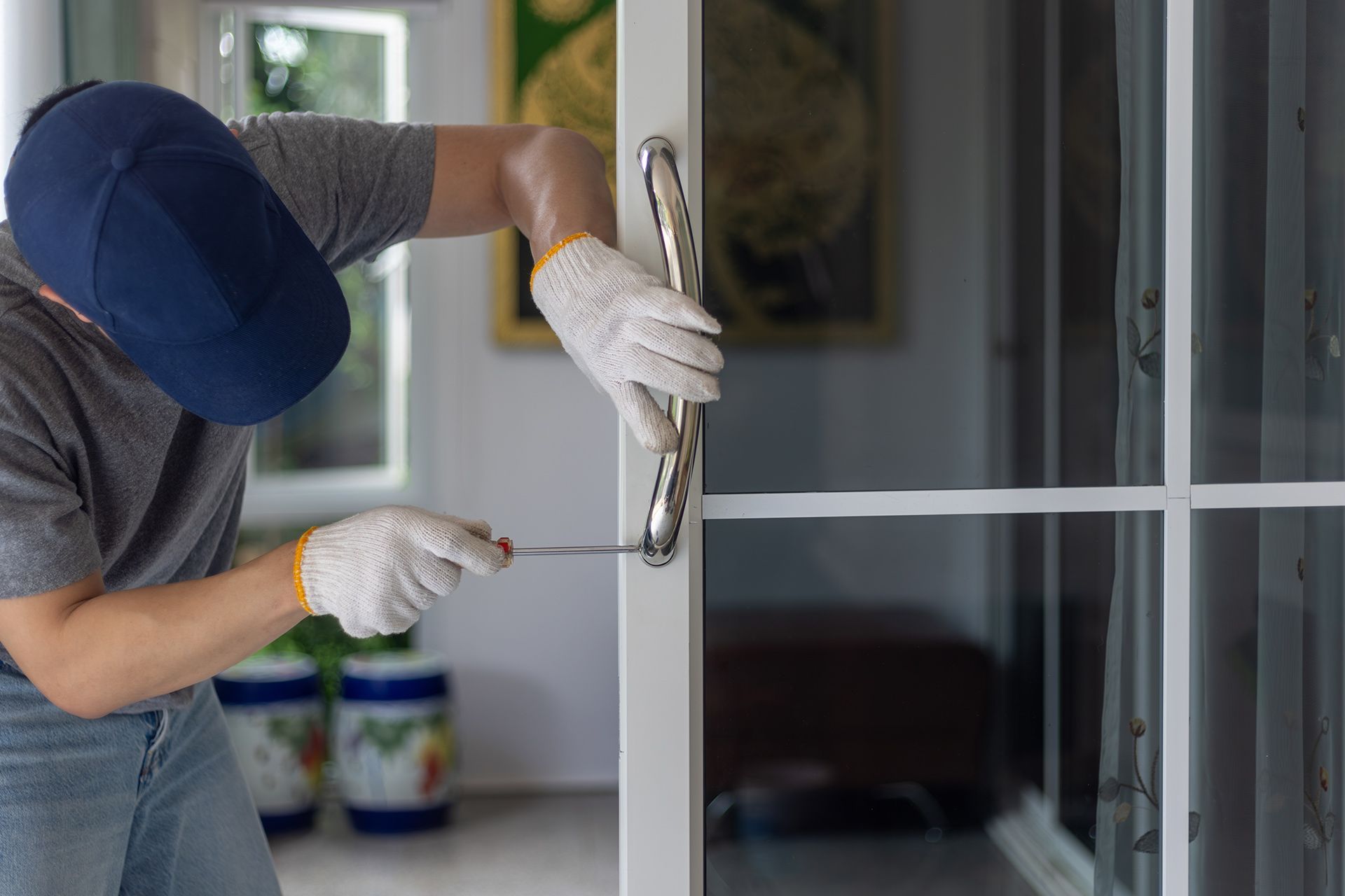 A person in a blue cap and work gloves uses a tool to repair or install a metal handle on a white glass door.