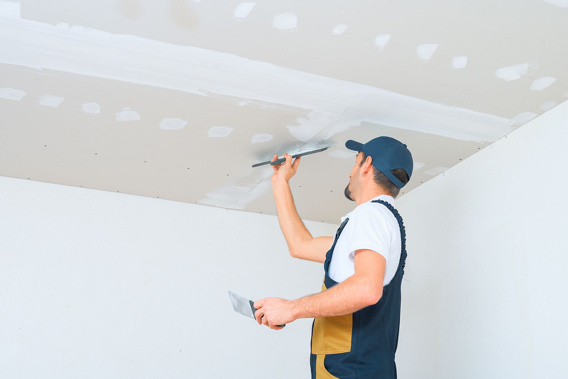 A worker wearing a blue cap and work overalls applies joint compound to a ceiling with a metal trowel.