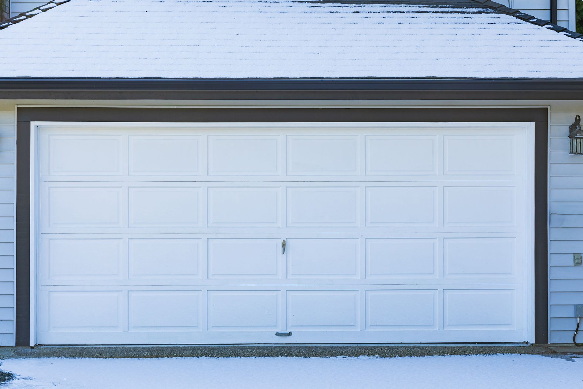 A white, four-panel residential garage door with brown trim, set in a house wall with snow on the roof and ground.