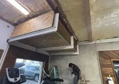 A person on a ladder working on the ceiling. Garage with treadmill, truck visible through window.