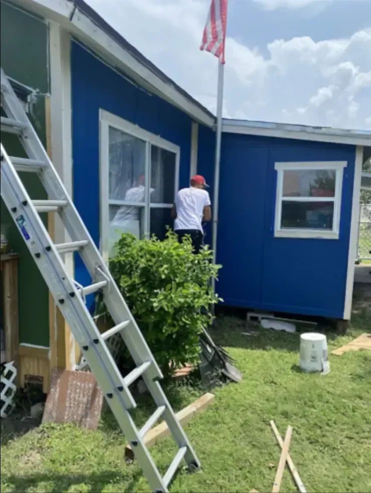 Man installing window in blue building. Silver ladder and US flag pole nearby.