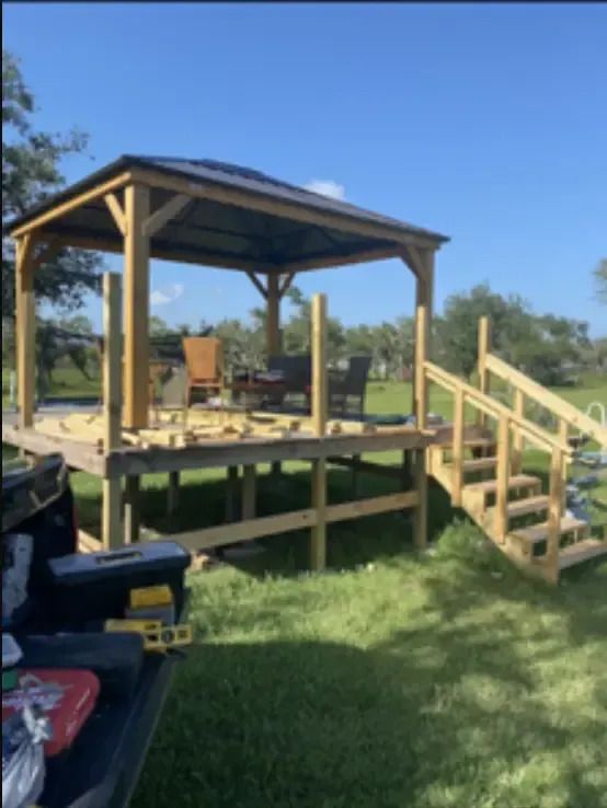 Wooden gazebo on a raised deck with stairs, under construction, set in a grassy yard under a blue sky.