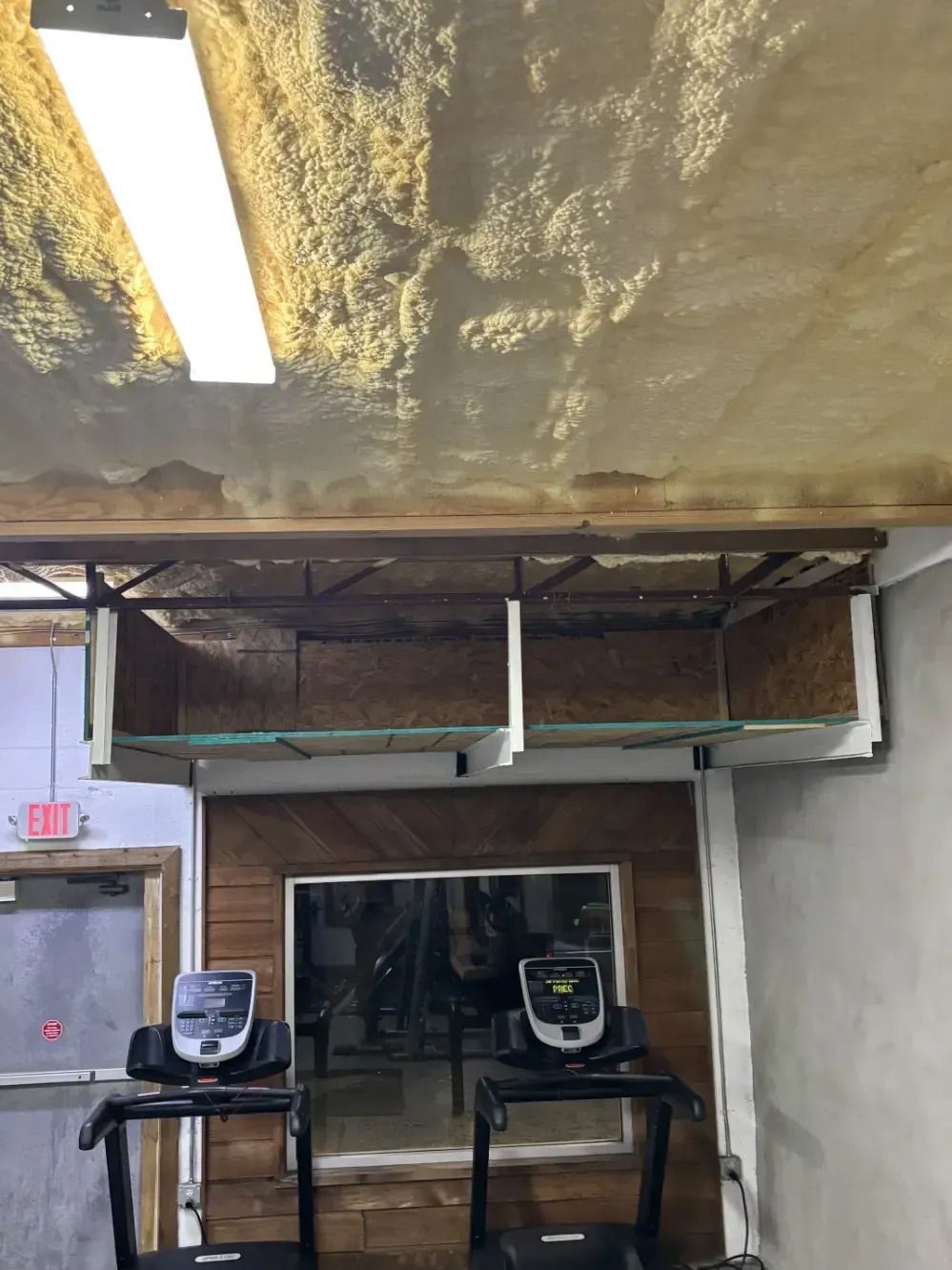 Two treadmills below a TV, set against a wood-paneled wall. A shelf above. The ceiling has stains.