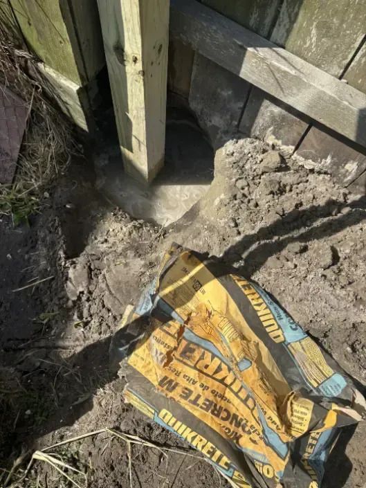 A wooden fence post set in concrete, next to a partially visible fence and a bag of concrete mix.