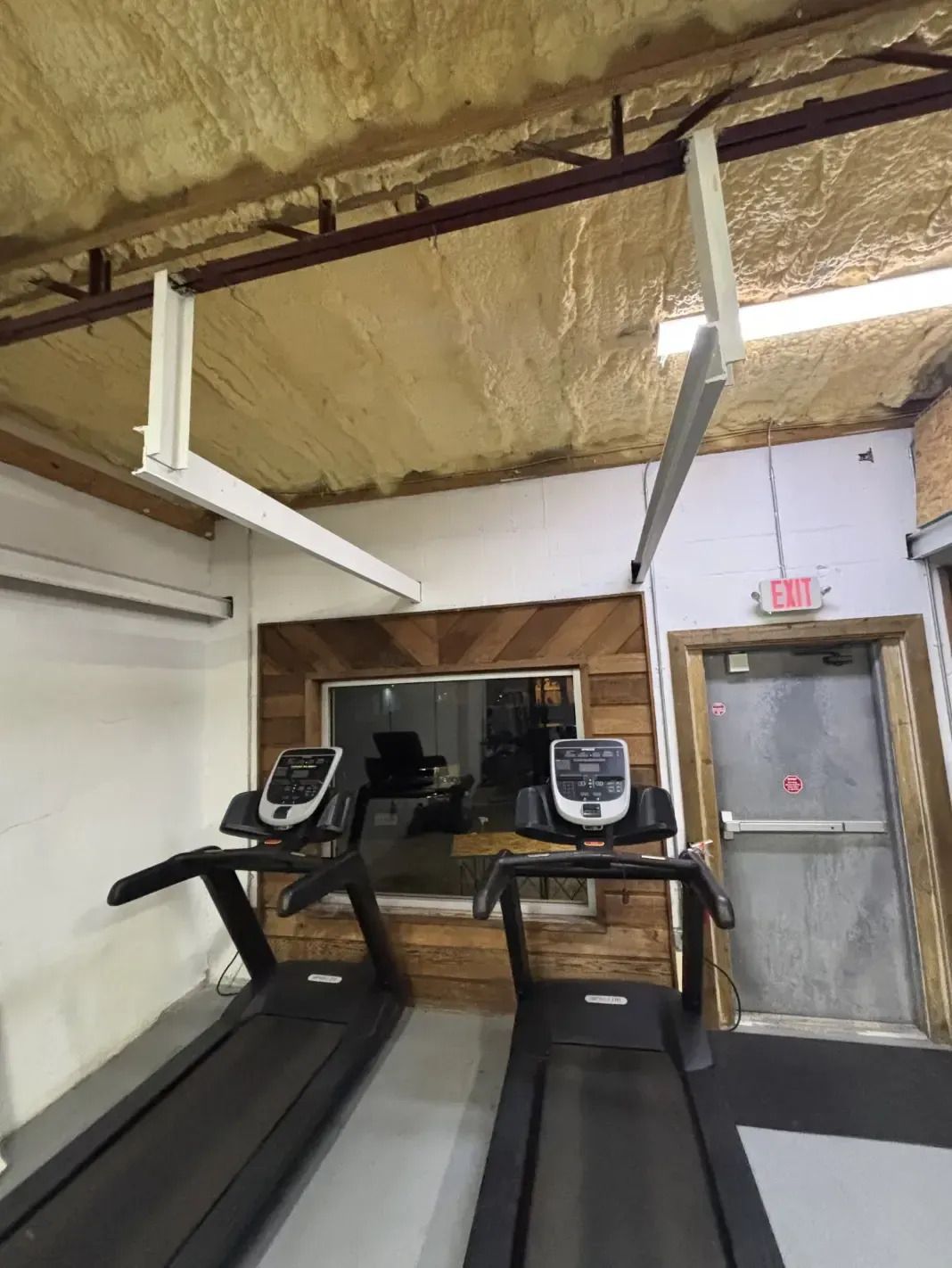 Two treadmills in a gym; white ceiling-mounted fans, window with wood paneling, exit door.