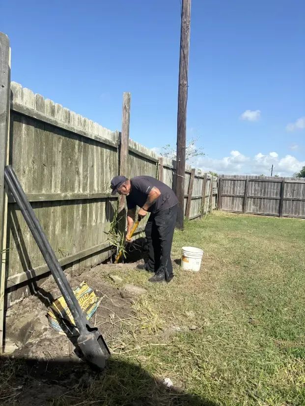 Person digging in yard next to a wooden fence on a sunny day. A shovel and bucket are nearby.