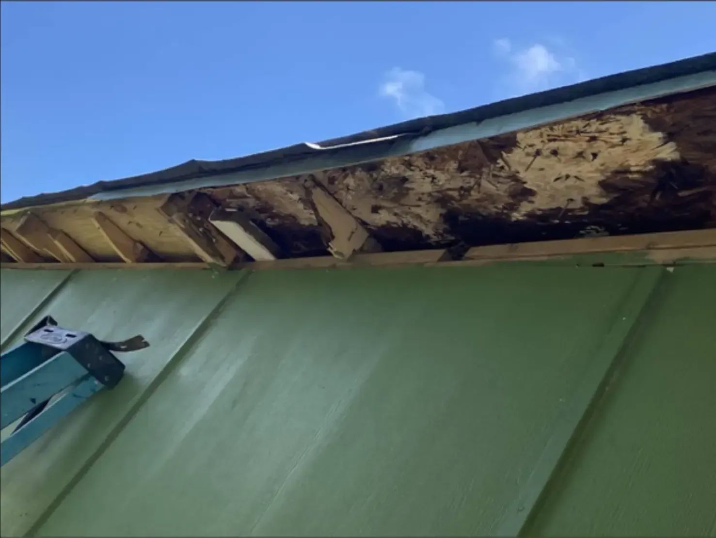 Damaged roof edge with rotten wood; green building exterior, blue sky.