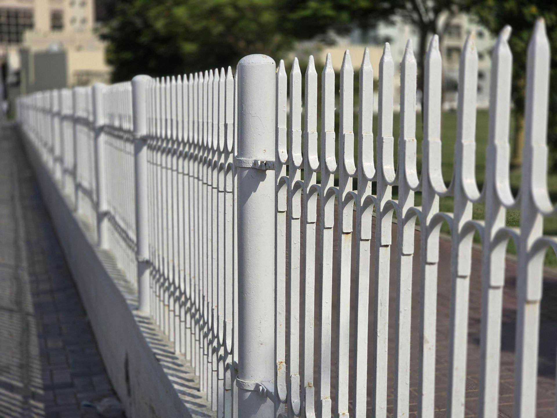 A white metal fence with pointed pickets lines a sidewalk in an outdoor setting.