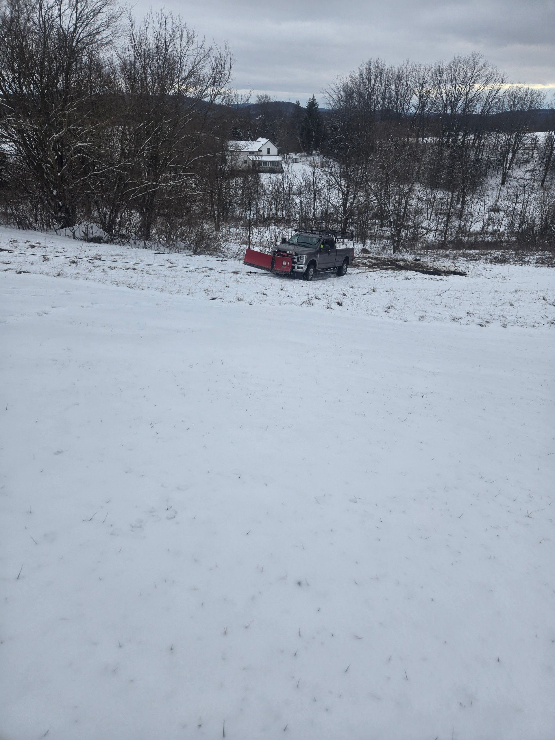 Snow-covered field with a truck partially buried in snow. Trees and a house are visible in the distance under a cloudy sky.
