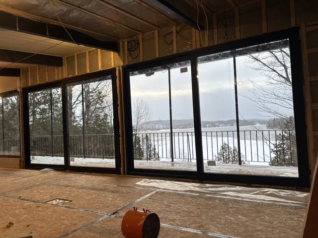 De grandes fenêtres offrent une vue sur un lac enneigé et des arbres. Les encadrements sont noirs. L'intérieur est inachevé.