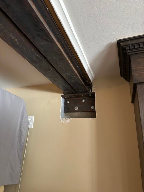 Poutre en bois foncé fixée sur un mur beige et reliée à un plafond blanc. Support métallique et vis apparents.