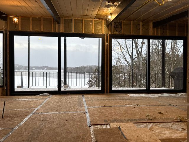 Vue intérieure d'une pièce aux grandes fenêtres à cadre noir donnant sur un paysage enneigé et un lac. Travaux en cours.