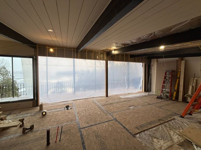 Intérieur d'une pièce en construction avec une bâche en plastique sur une fenêtre, des poutres apparentes et un sol non fini.