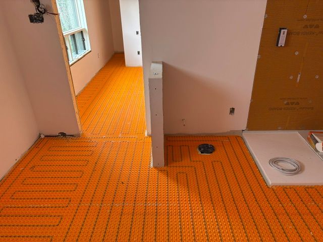 Un tapis chauffant orange au sol est installé dans une pièce. On aperçoit également une porte, des murs blancs et un receveur de douche.