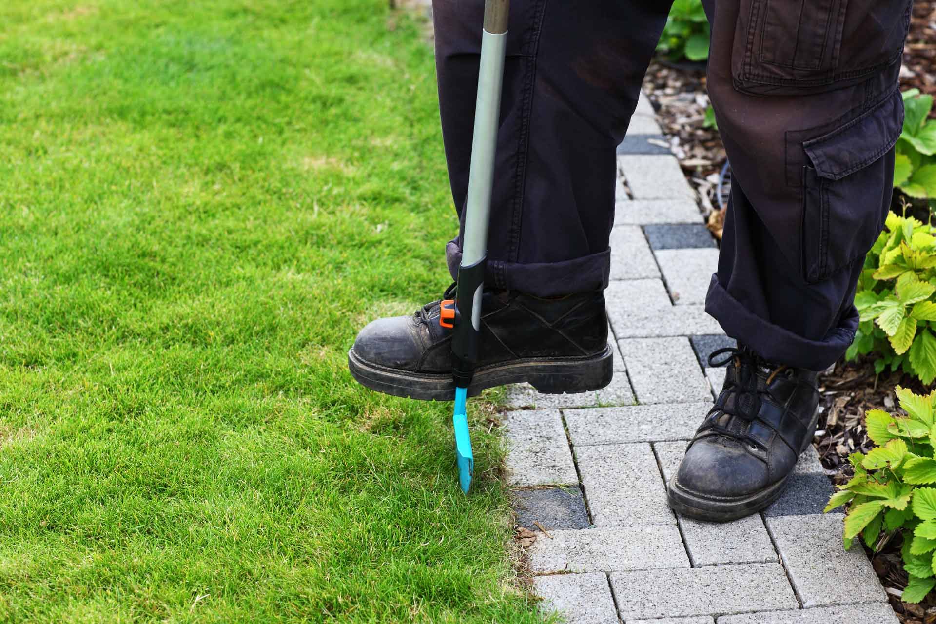 A person is standing on a sidewalk cutting grass with a lawn mower