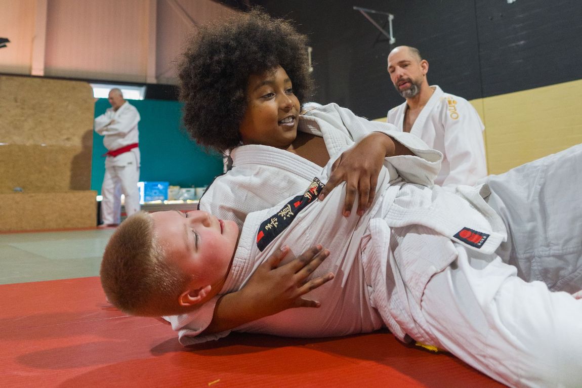 Twee mensen in witte judopakken oefenen een houdgreep  op een rode mat in een sportschool, terwijl een instructeur van dichtbij toekijkt.