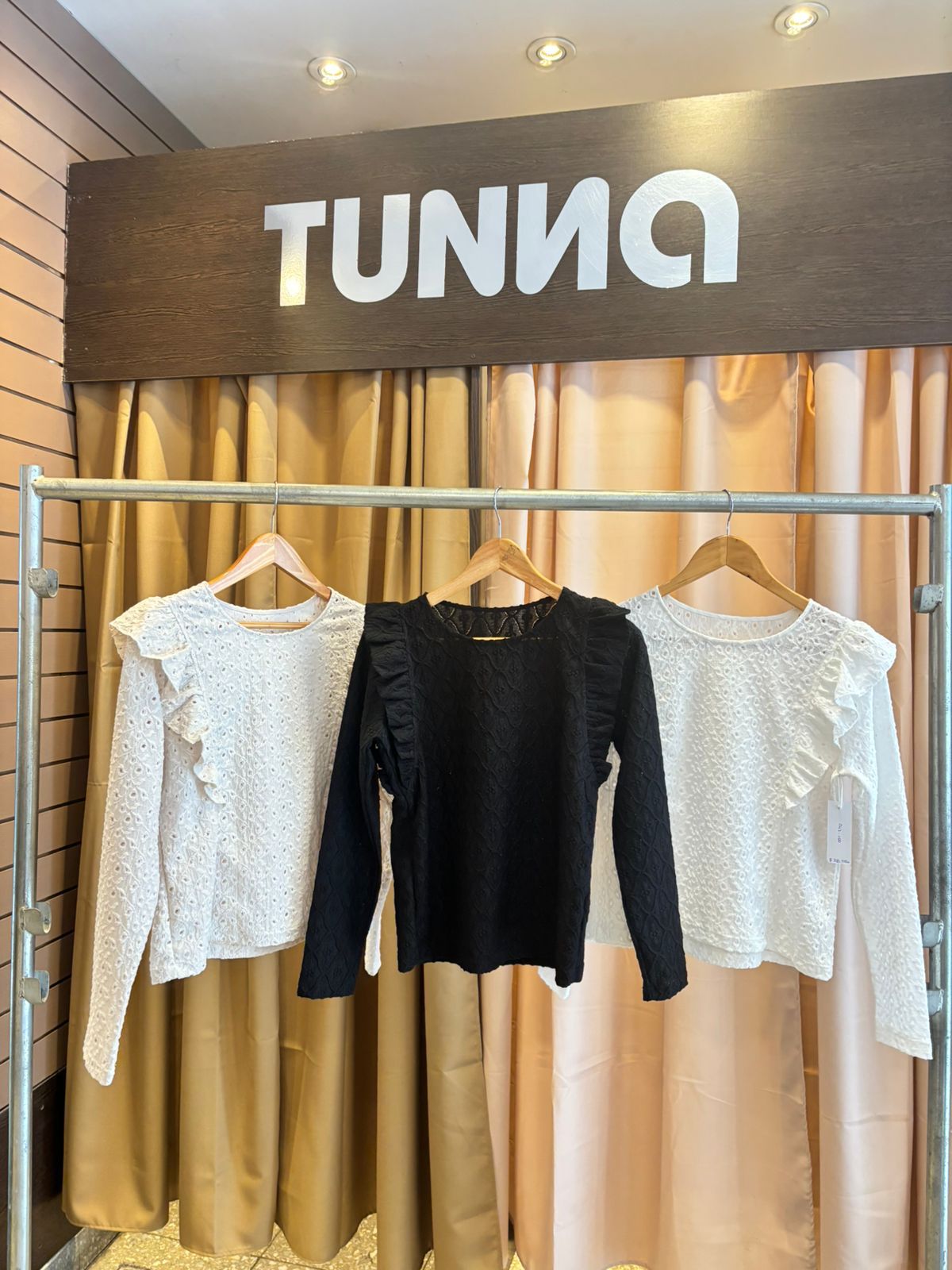 Tres camisas de manga larga colgadas en un perchero. Dos blancas y una negra, todas con volados