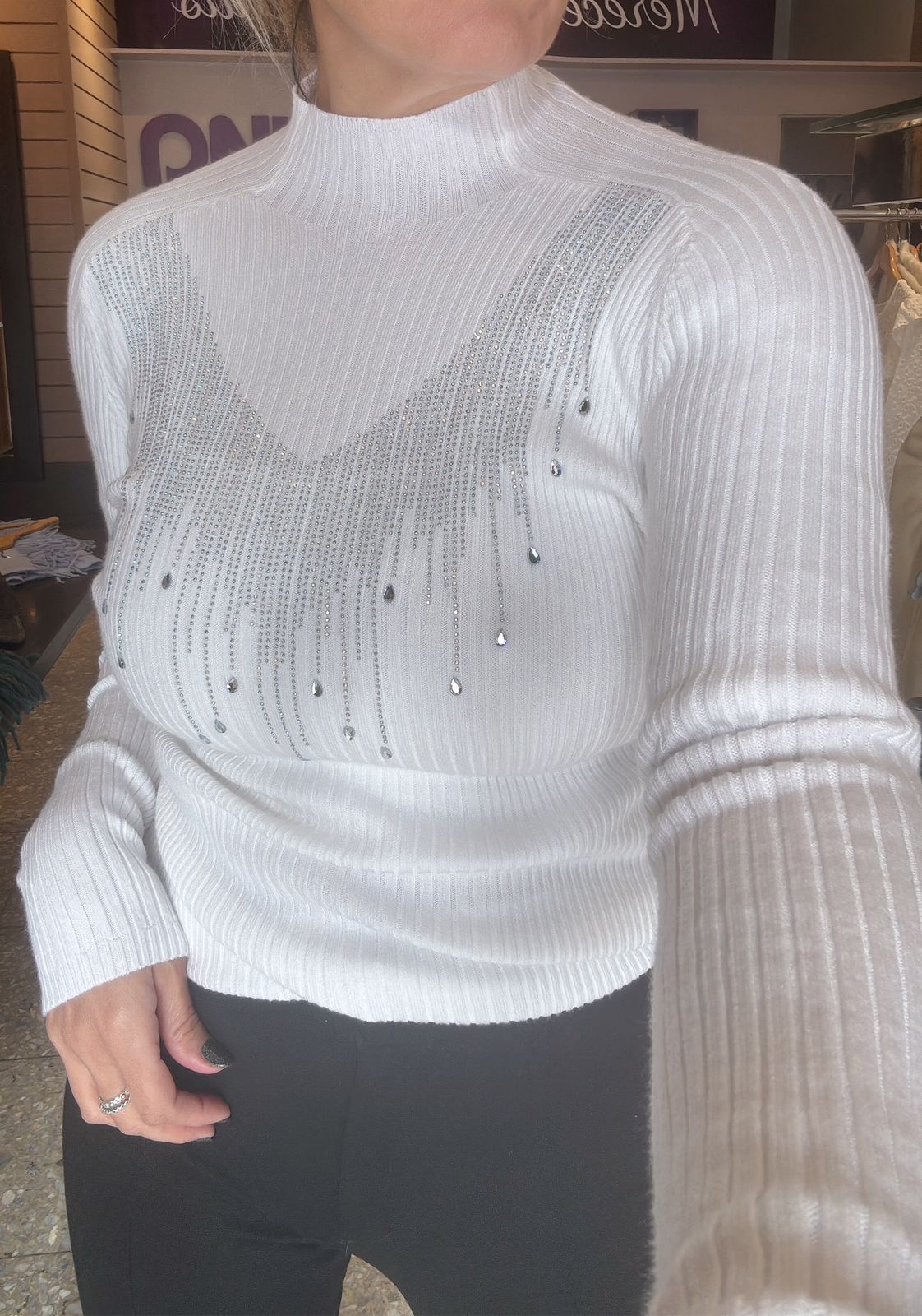 Mujer con suéter color canela con adornos, pantalón negro, sonriendo, en un entorno boutique.