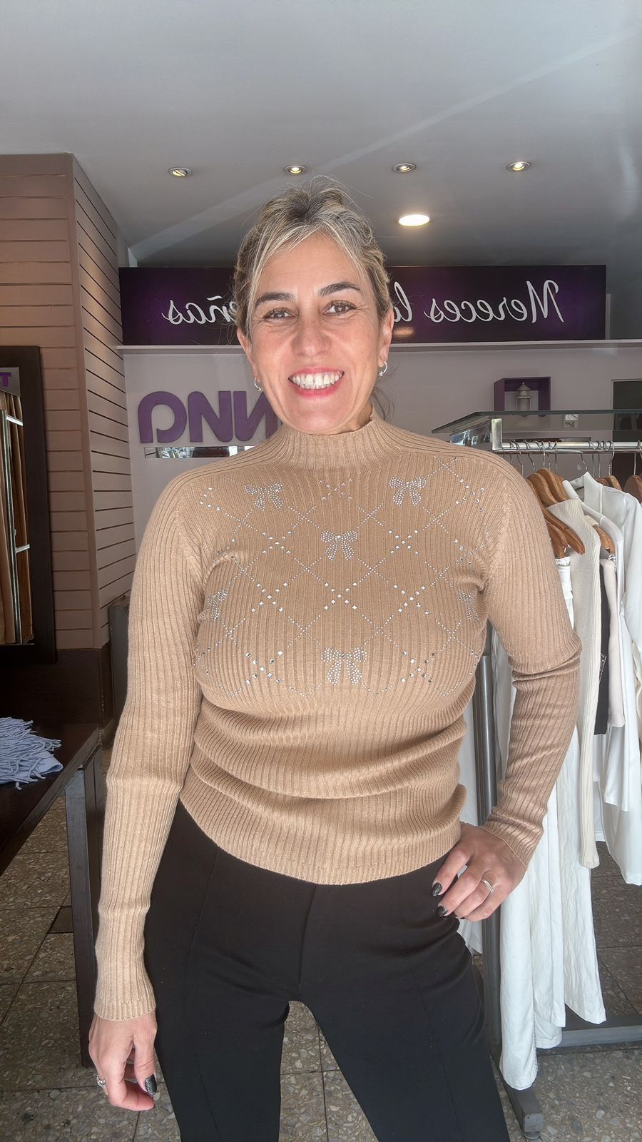 Mujer con suéter color canela con adornos, pantalón negro, sonriendo, en un entorno boutique.