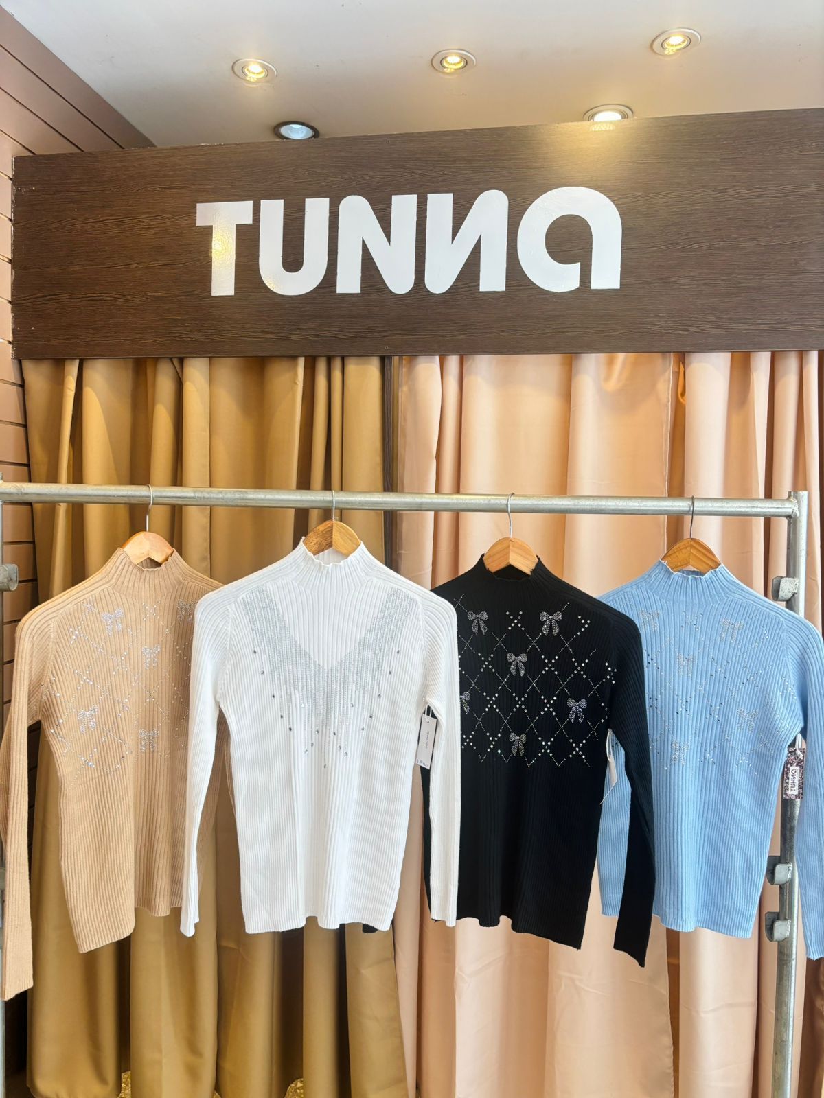 Cuatro suéteres colgados en un perchero: beige, blanco, negro (con estampado de lazos) y azul claro. Letrero "TUNNA" arriba.