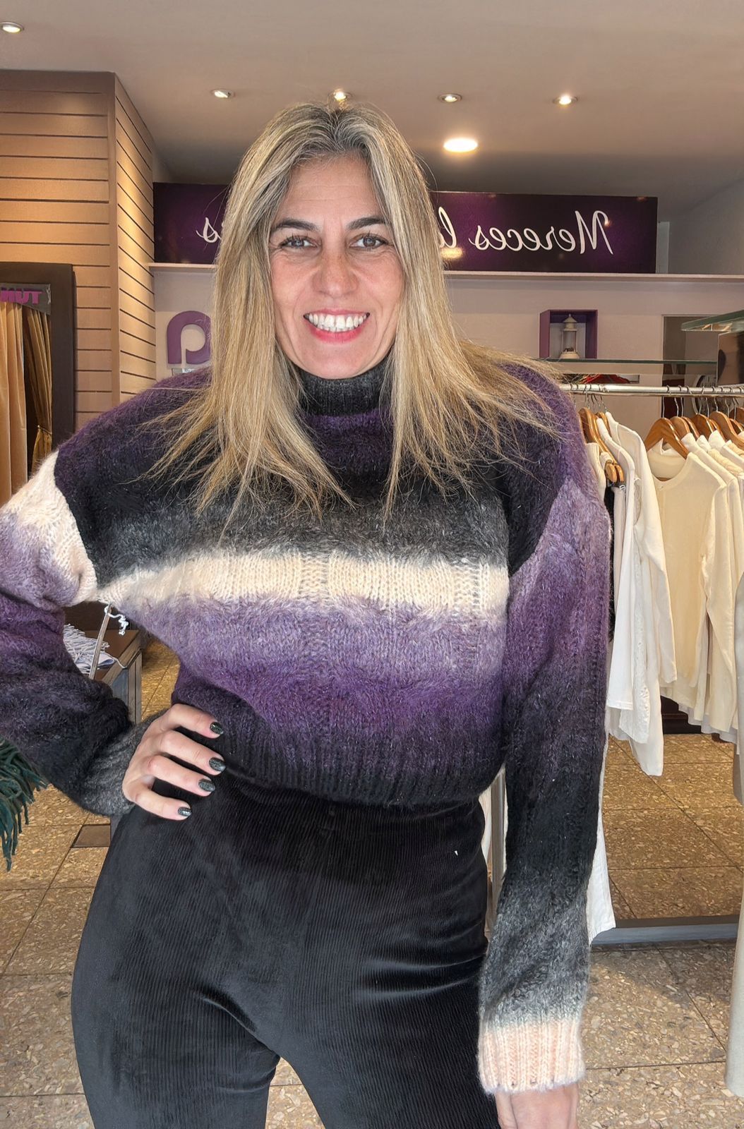 Mujer sonriendo, vistiendo un suéter degradado morado y negro y pantalones negros en una tienda de ropa.