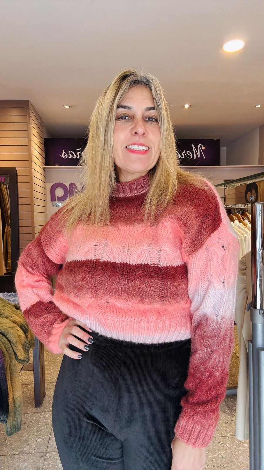 Mujer sonriendo, vistiendo un suéter degradado morado y negro y pantalones negros en una tienda de ropa.