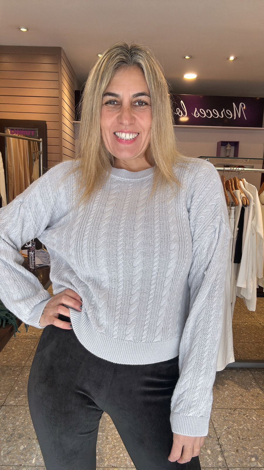 Una mujer con un suéter gris claro y pantalones negros sonríe en una tienda de ropa.