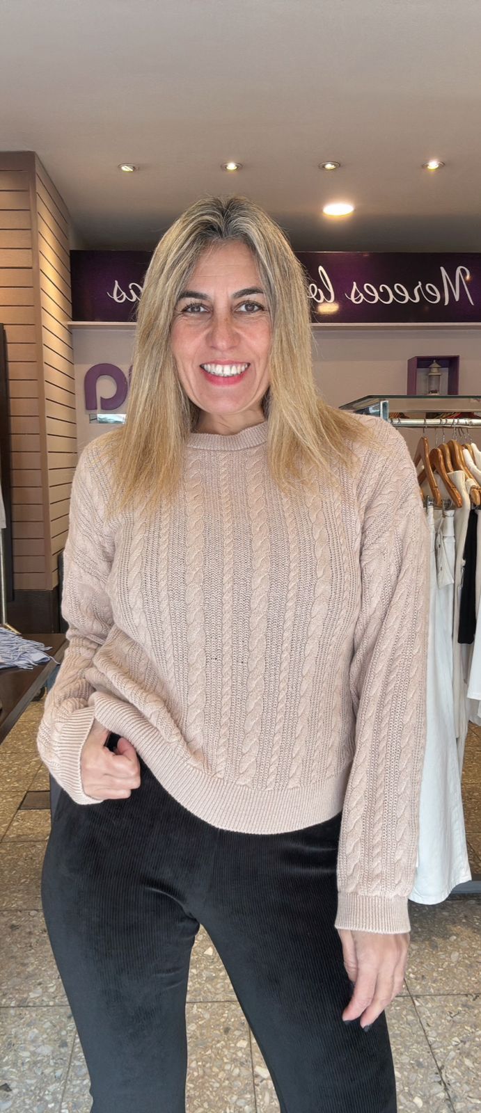 Una mujer con un suéter beige y pantalones negros sonríe, parada en una tienda.