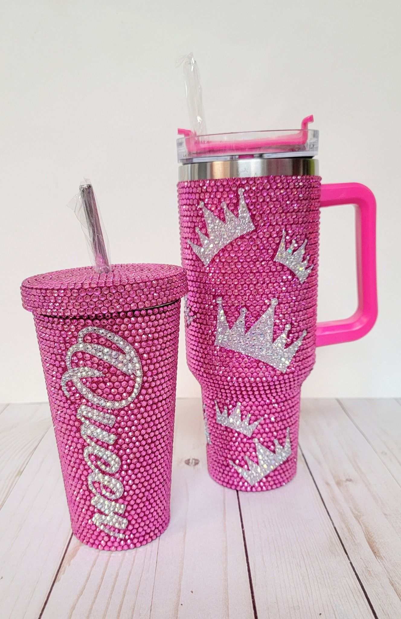 Two bedazzled pink tumblers with crowns; one with a name, and one with a handle and straw.