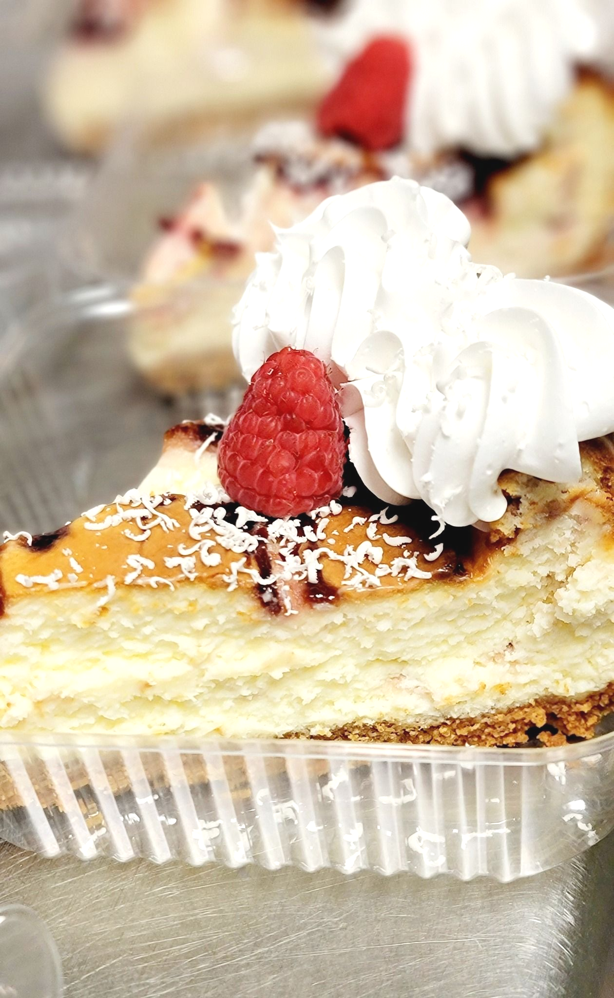 Cheesecake slice in a plastic container, topped with whipped cream, raspberry, and chocolate drizzle.