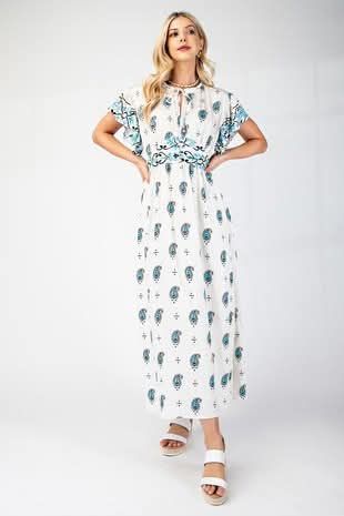 Woman in white patterned maxi dress and sandals, hands on hips, standing against a white background.