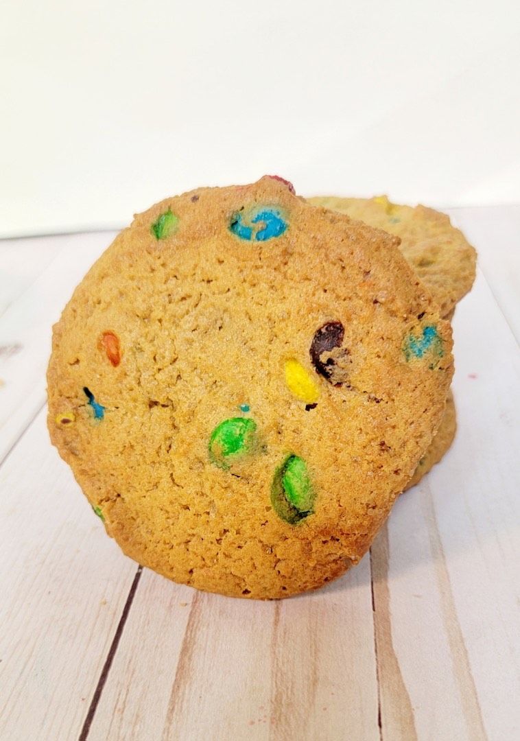 Two M&M cookies on a white wood surface. The cookies are brown and have colorful candies.