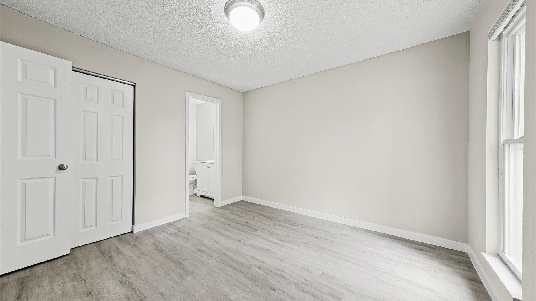 Empty neutral bedroom with white closet doors, a window on the right, and an open doorway to a bathroom.